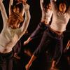 Dancers in white long sleeves and black pants with their arms raised over their head with their focus looking over their shoulder