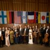 Musicians hold their instruments while standing in a line beneath various international flags