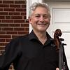 Man holding a cello in front of a brick and white wall