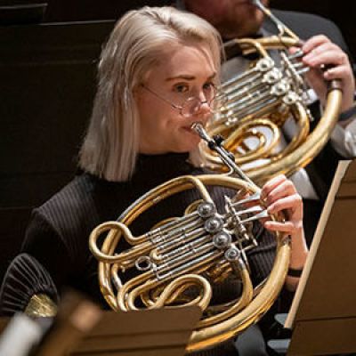 Photo of young woman with blonde hair playing the French horn.