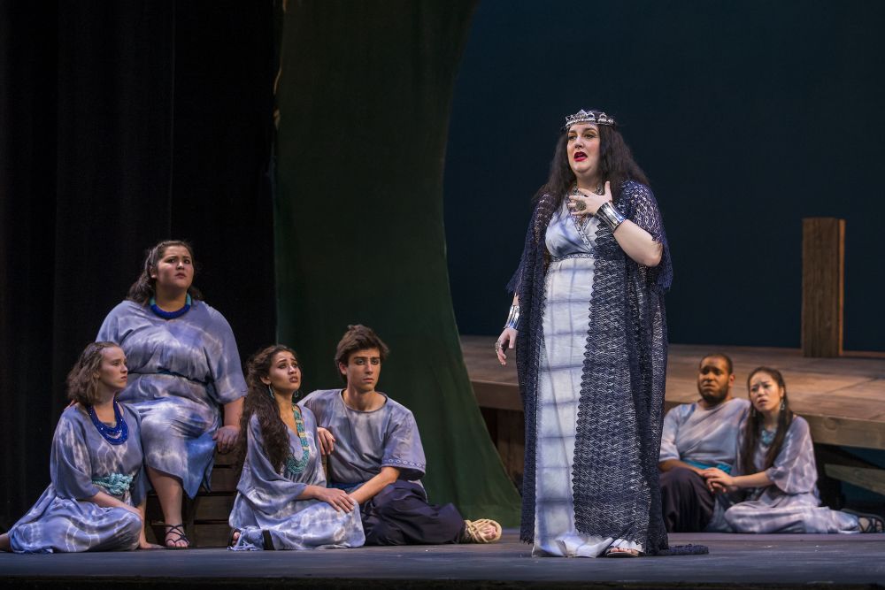 Photo of Dido and Aeneas being performed on stage. Woman in blue dress and crown standing and singing while ensemble of students dressed in blue tie-dyed costumes watch