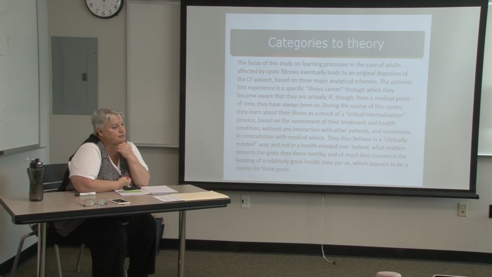 Photo of professor sitting at desk beside a pull-down screen with slide that says "Categories to theory" with some text underneath