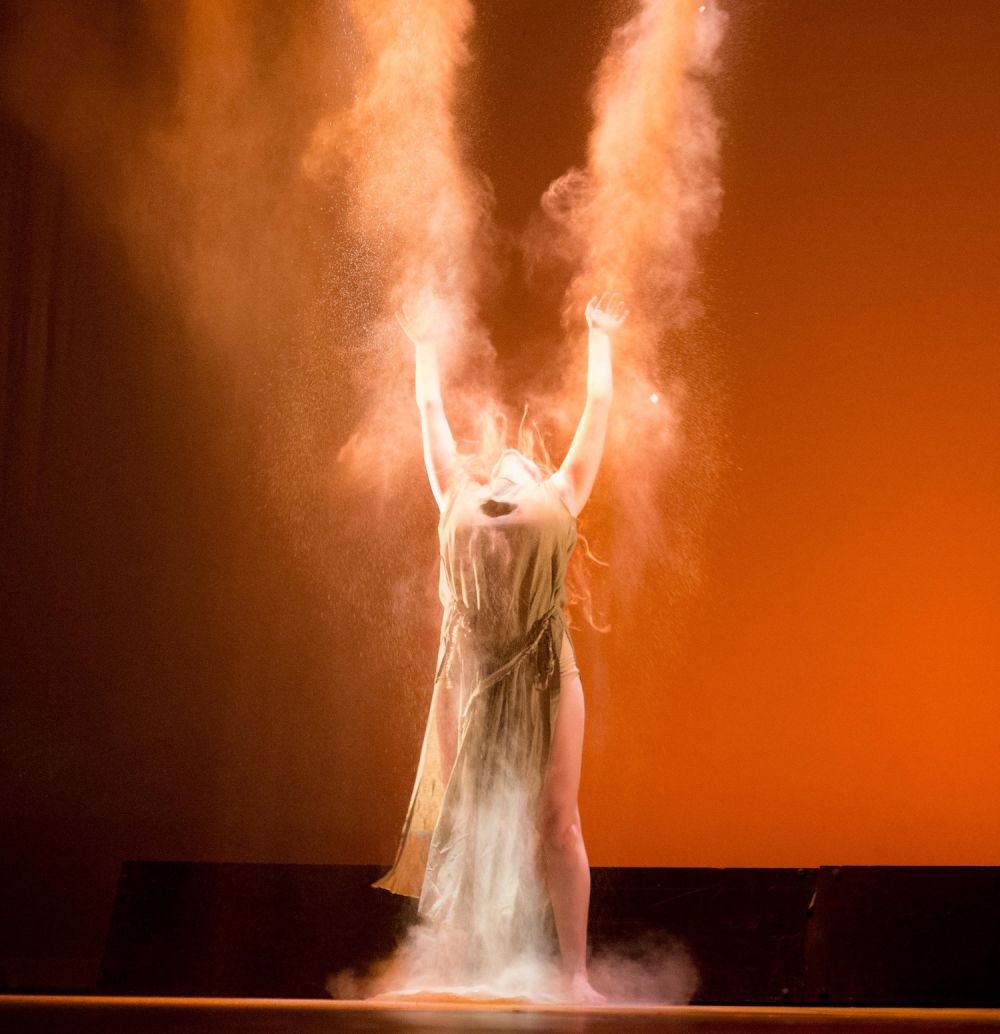 Performer with arms raised above head against an orange backdrop.