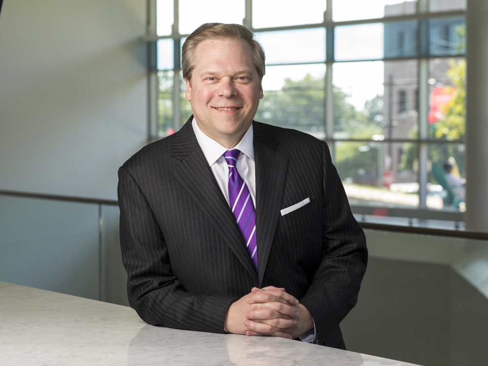 Man with gray hair wearing pinstripe suit and purple striped tie, resting forearm on ledge