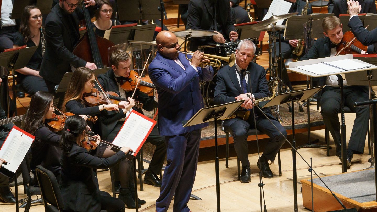 Trumpet soloist performs with symphony orchestra and jazz band surrounding him on stage.